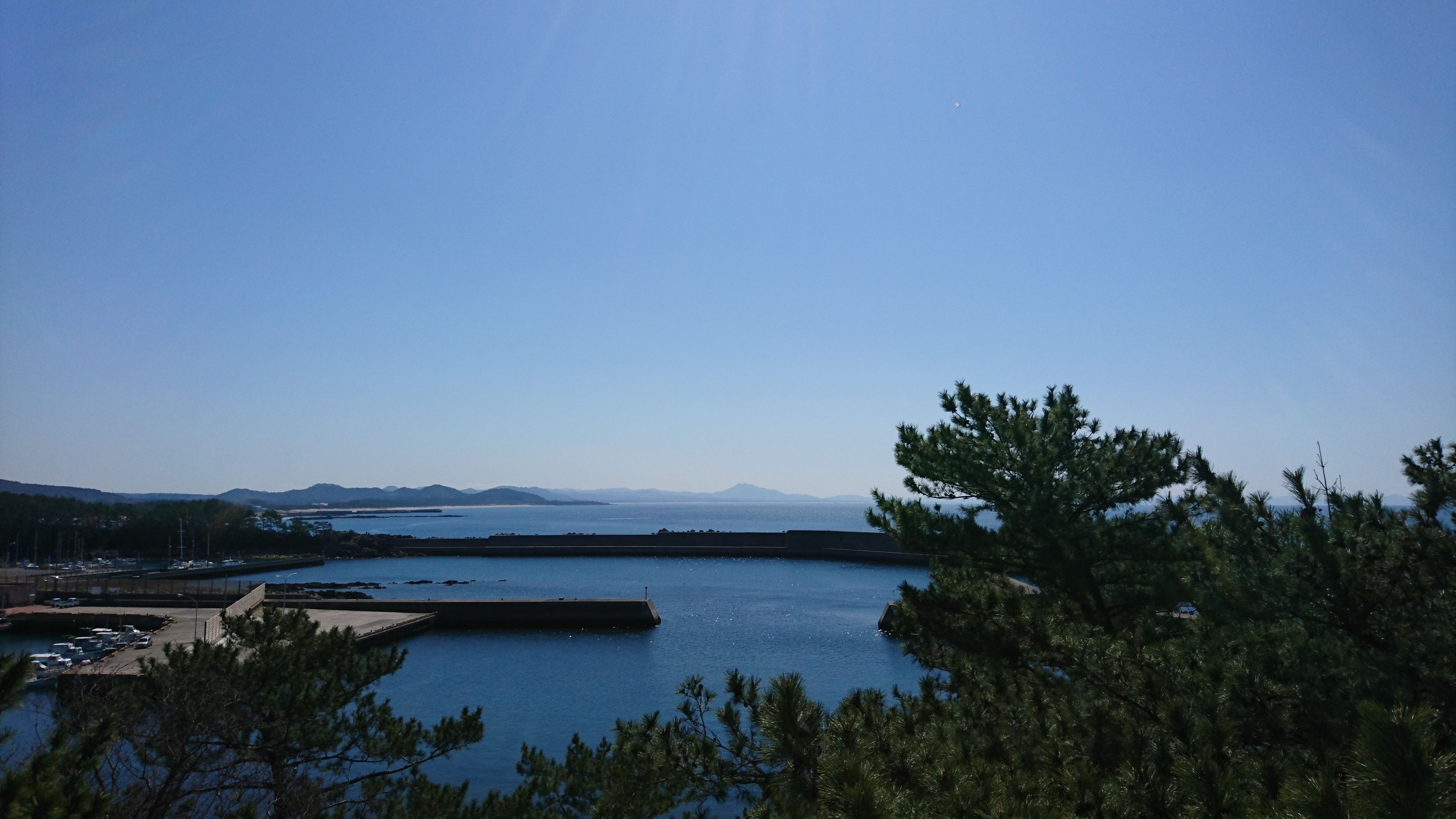 Vista costera con un puerto deportivo, cielo despejado y montañas a lo lejos.