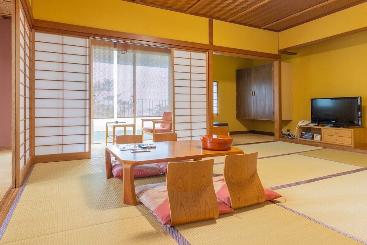 Una habitación japonesa tradicional con suelo de tatami, una puerta corredera, una mesa con cojines y un televisor.