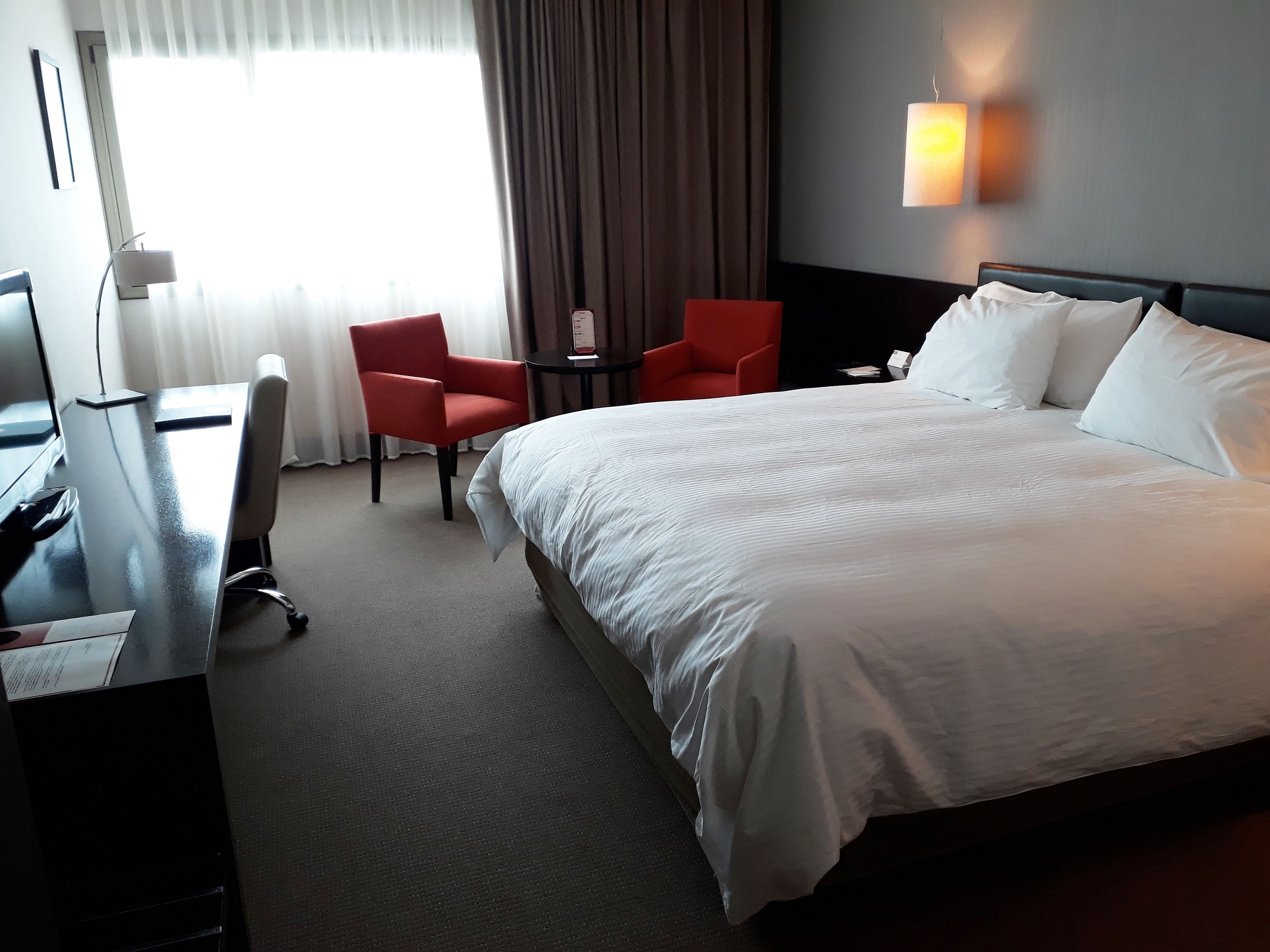 A hotel room with a large bed, two red chairs, a desk with a computer, and a lamp.