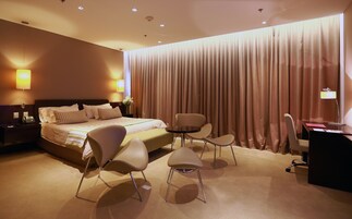 Quarto de hotel moderno com uma cama grande, duas poltronas, uma mesinha e uma escrivaninha com abajur.