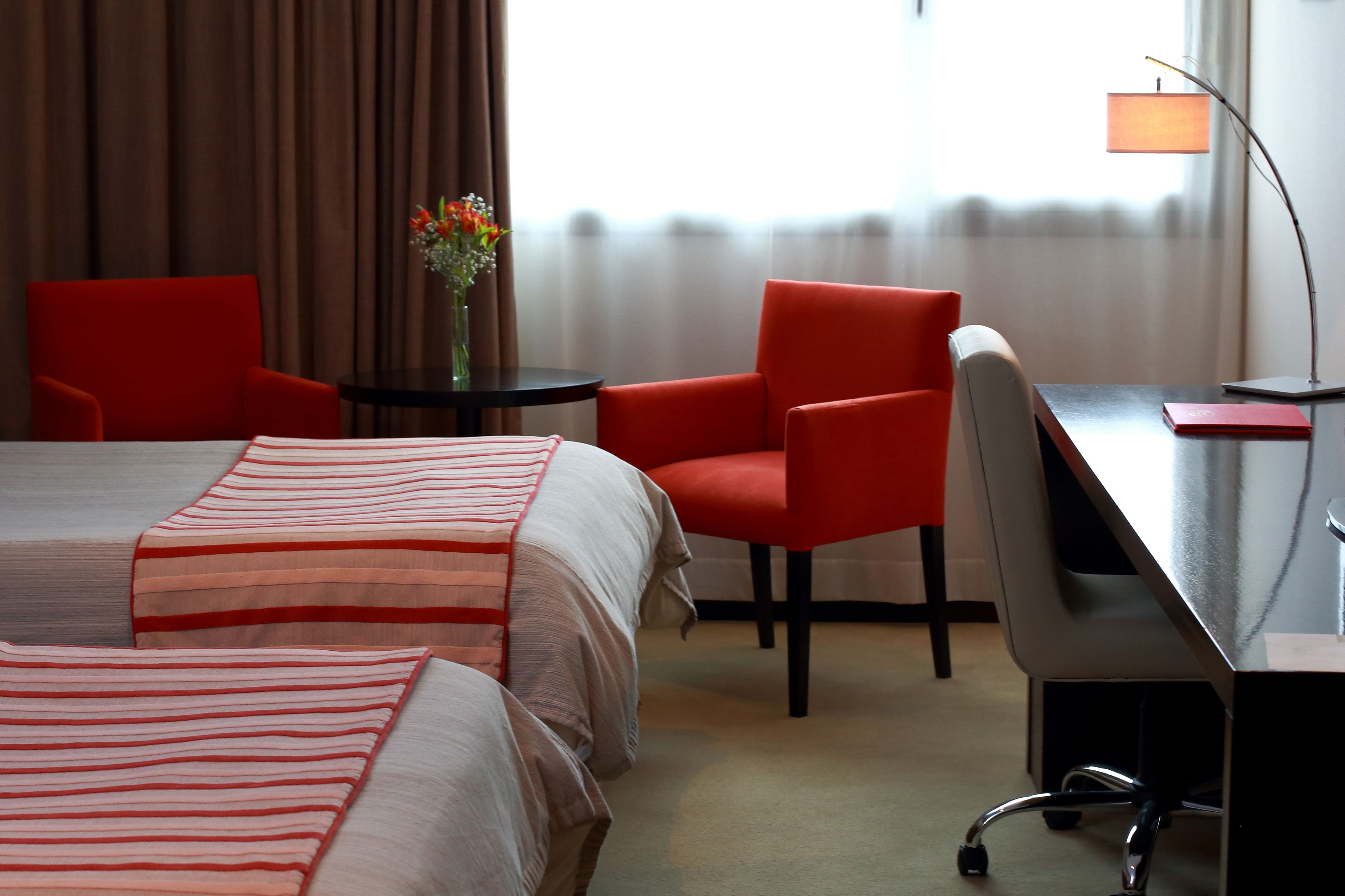 Et hotelværelse med en seng, to røde stole, et skrivebord med en lampe og en vase med blomster.