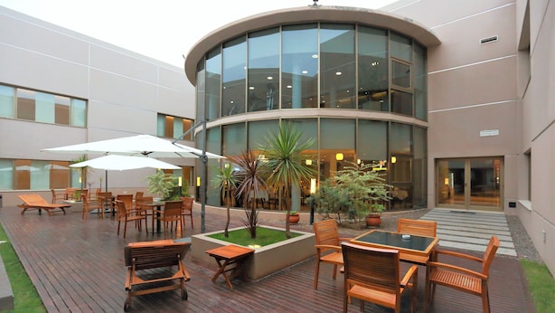Un patio exterior con mobiliario de madera, plantas en macetas y un edificio de vidrio al fondo.