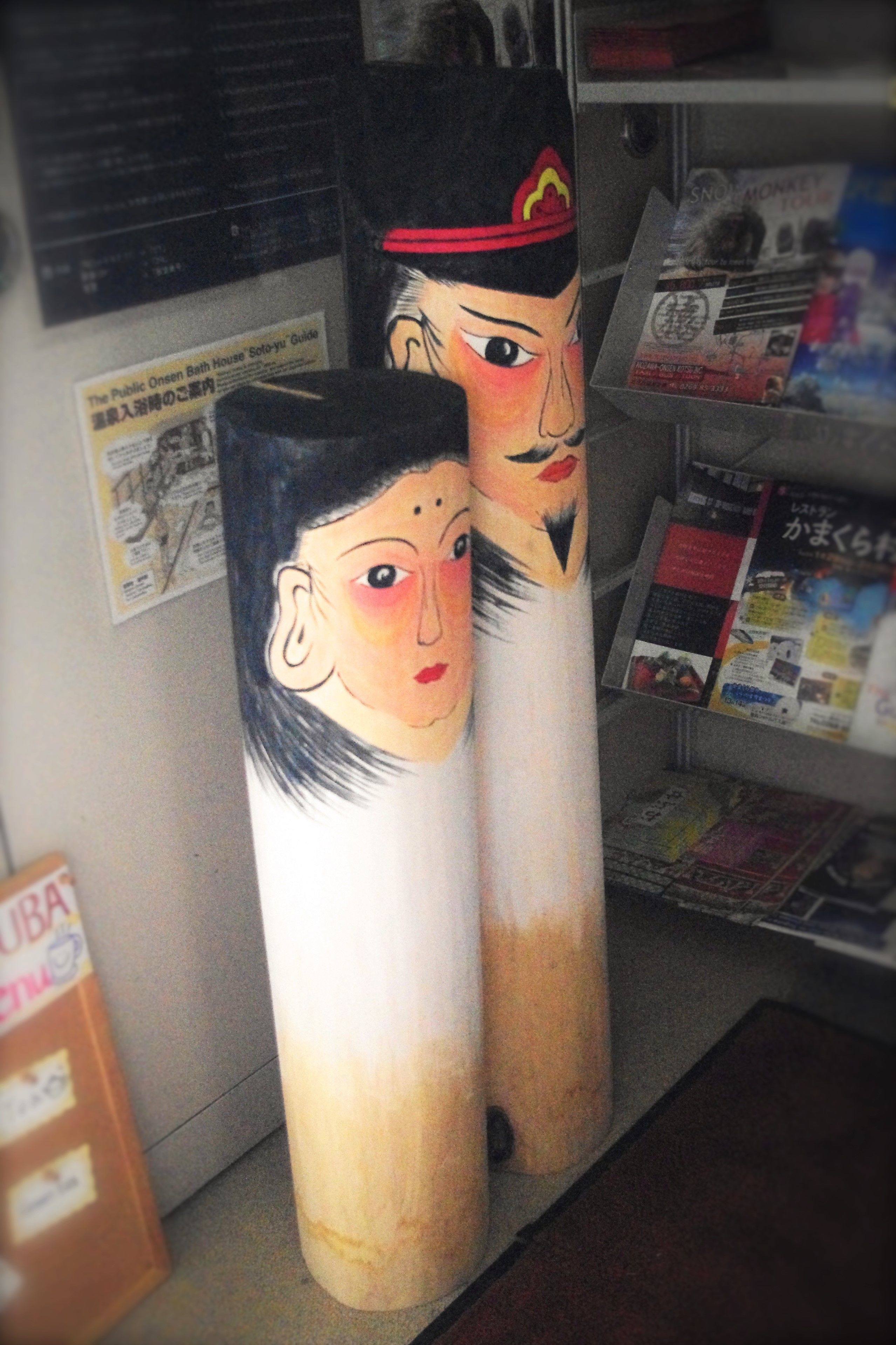 Two cylindrical objects with painted faces, a bulletin board with various papers, and a shelf with magazines.
