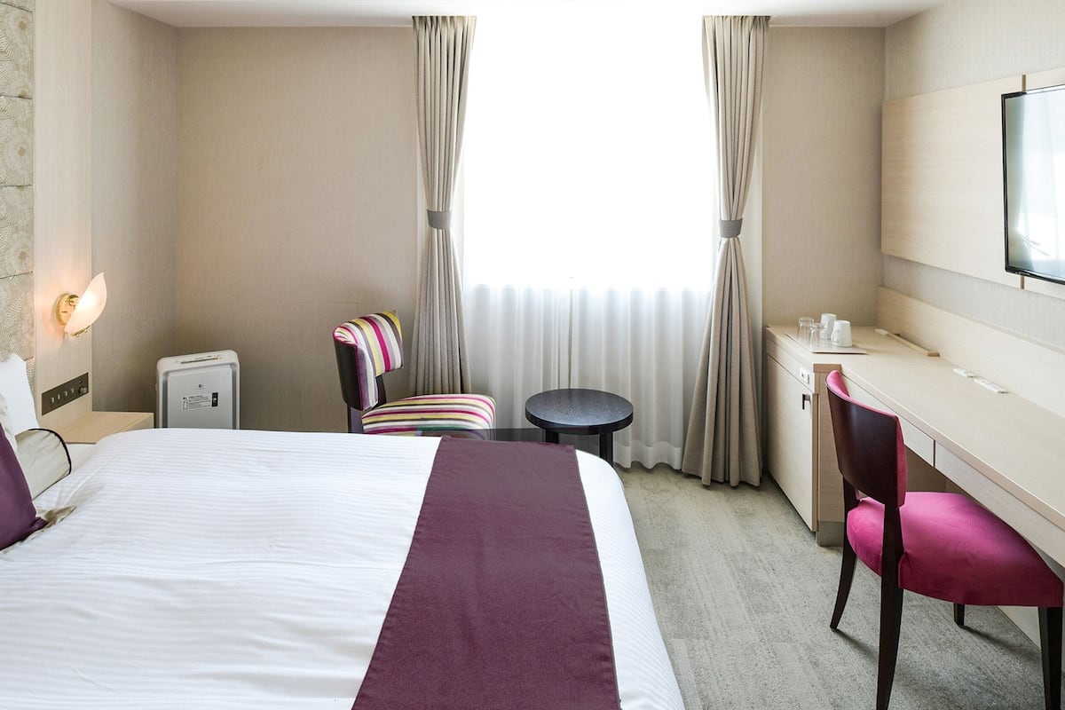 A hotel room with a bed, a desk, a chair, a TV, and a window with curtains.