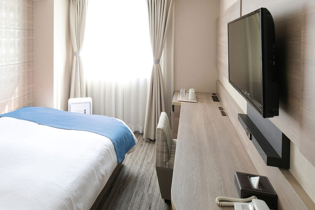 A hotel room with a bed, a TV, a bedside table, and a window with curtains.