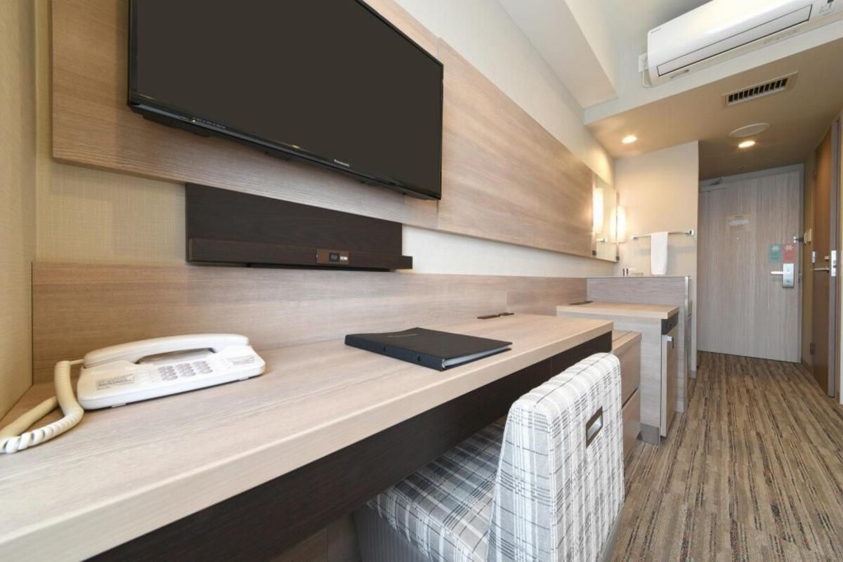 A hotel room with a flat-screen TV mounted on the wall, a desk with a telephone and a notebook, a chair, and a small kitchenette area.