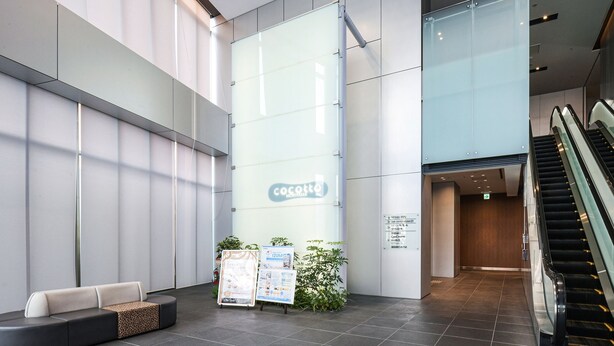A modern hotel lobby with a reception desk, a glass wall with signage, and an escalator. - Vessel Inn Shiga Moriyama Station (Moriyama)