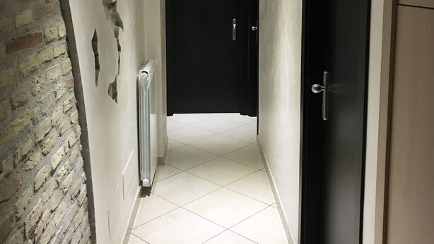 A hallway with a tiled floor, a brick wall on the left, and a black door at the end. - Sweety (Rome)