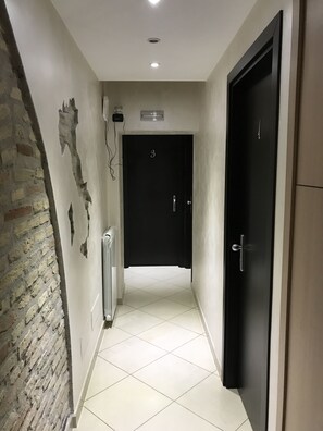 Un pasillo con suelo de baldosas, pared de ladrillo a la izquierda y una puerta negra al final.