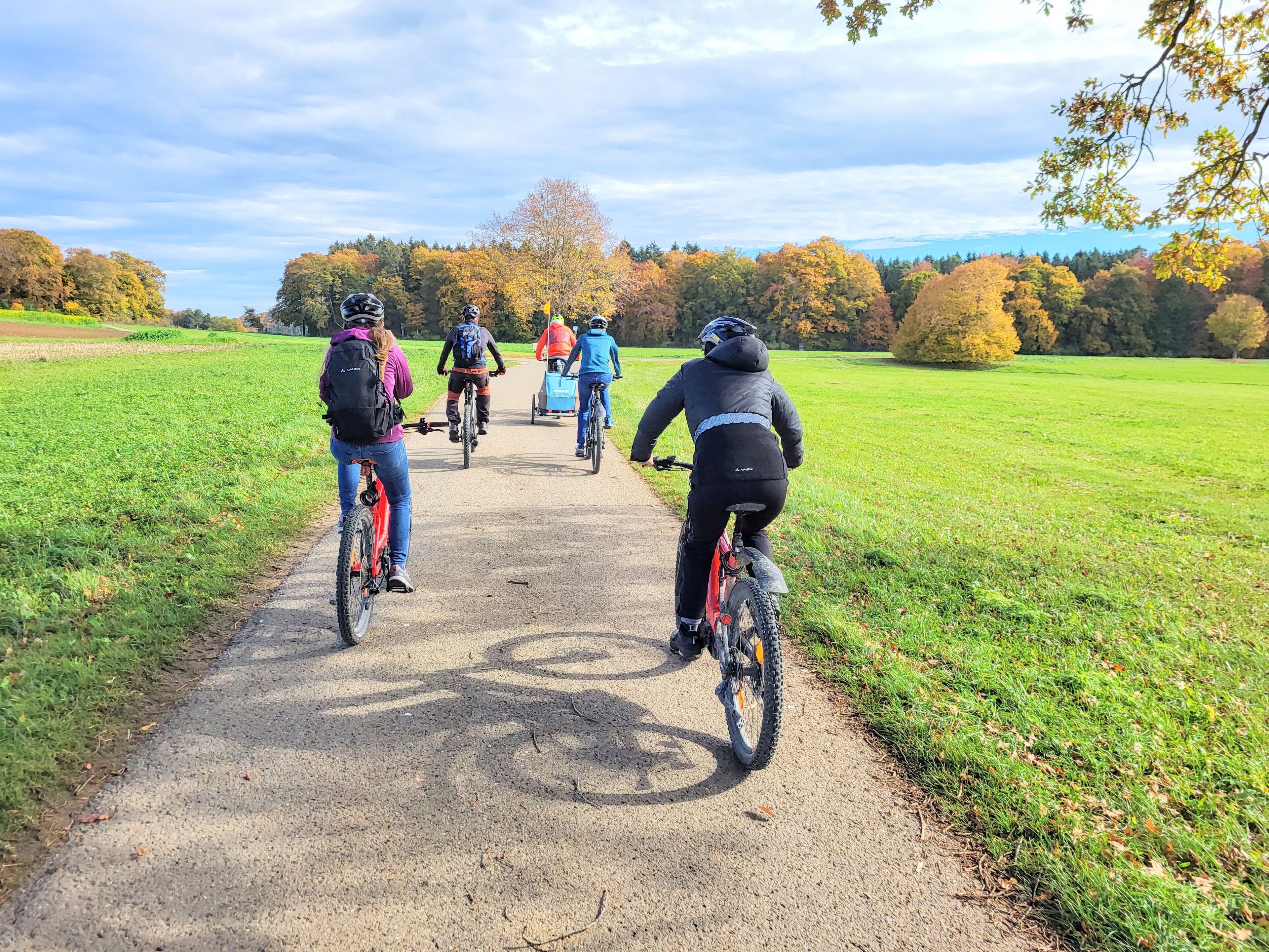 Um caminho pavimentado com ciclistas se afastando da câmera, cercado por campos verdes e árvores com folhagem de outono.