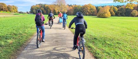 A paved path with cyclists riding away from the camera, surrounded by green fields and trees with autumn foliage.