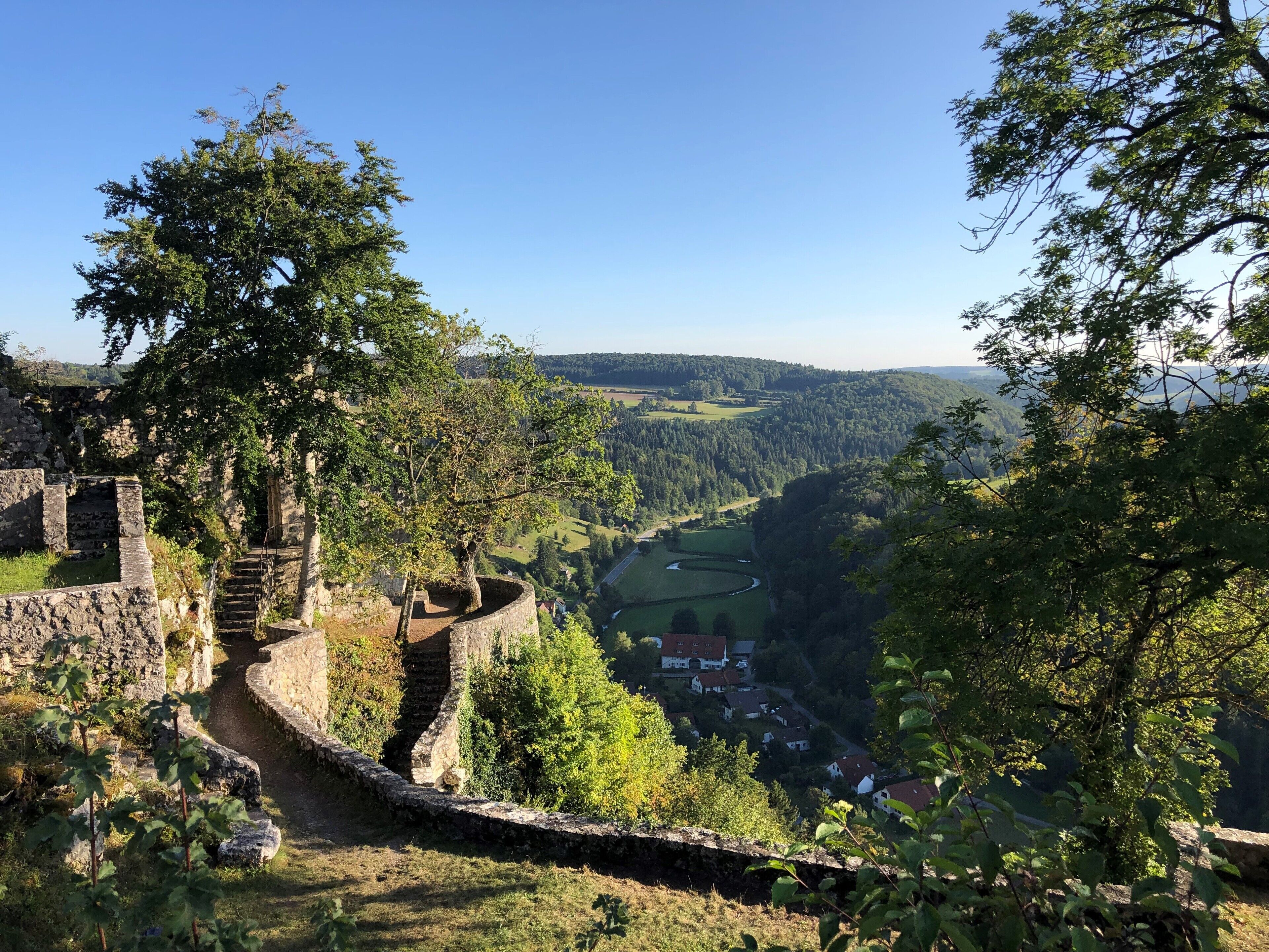 A historic stone wall with a staircase leading to a higher vantage point, overlooking a valley with houses and a river.