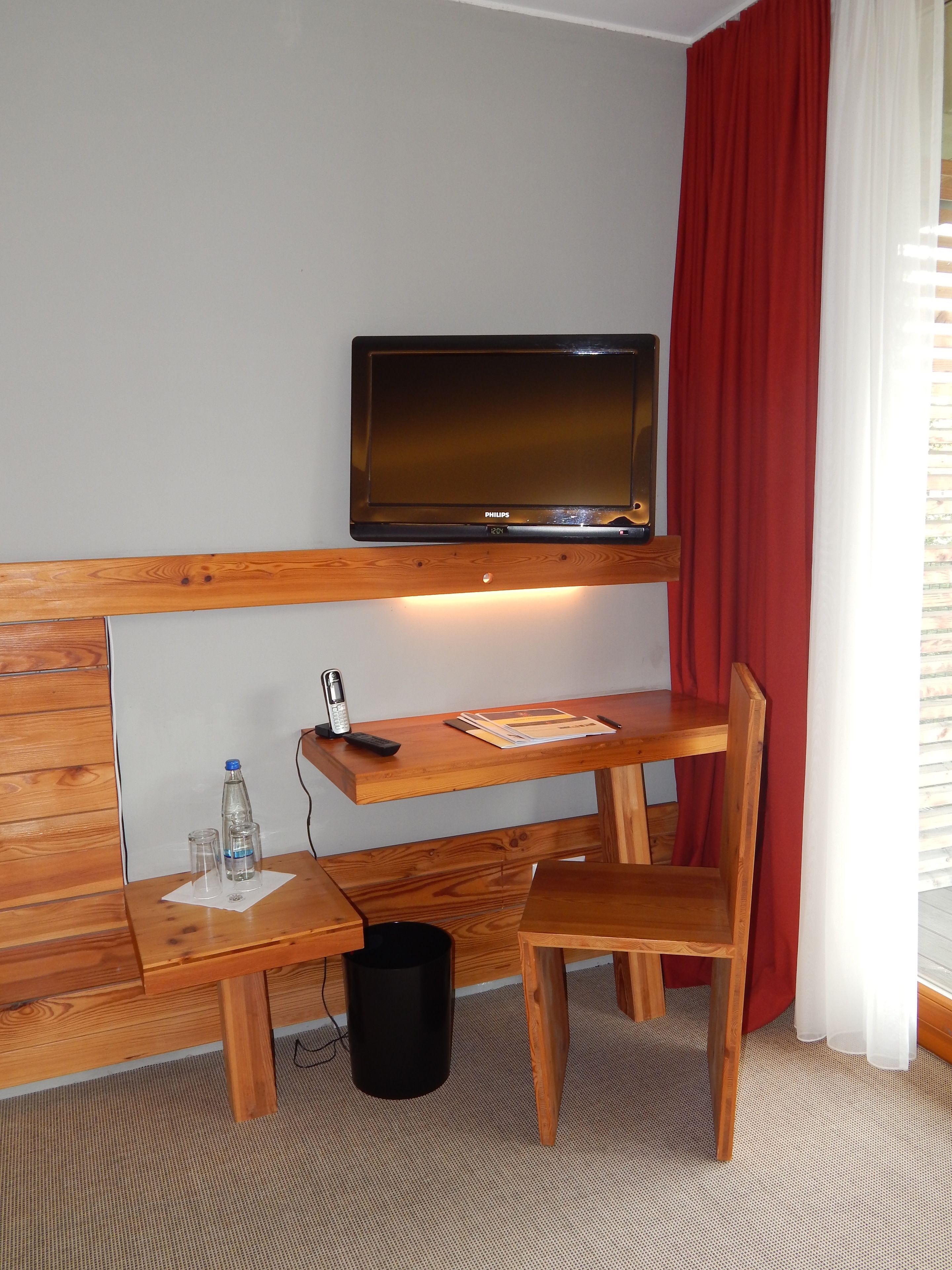 Ein Hotelzimmer mit einem Holztisch, einem an der Wand montierten Fernseher, einem roten Vorhang und einem Fenster mit Jalousien.