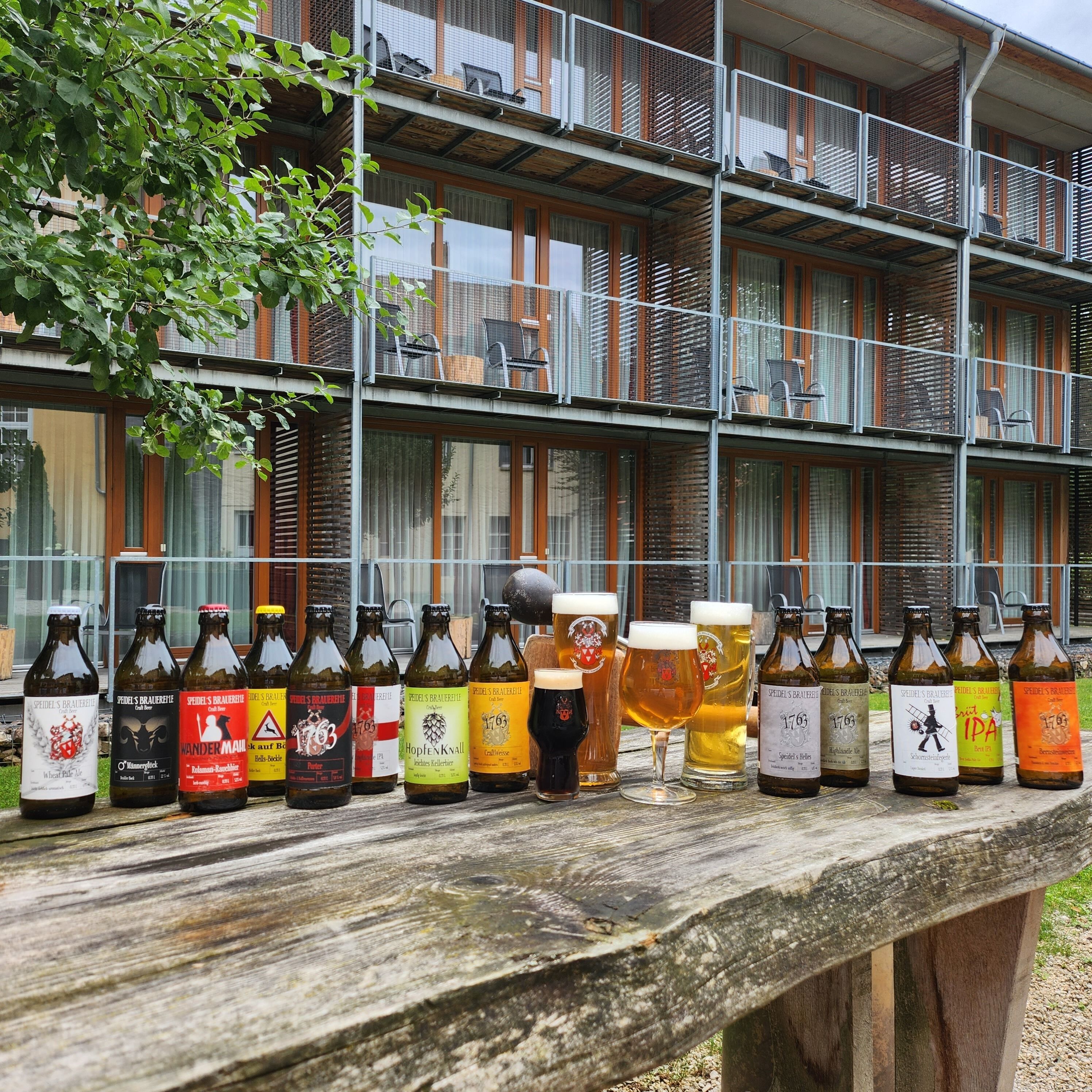 Een rij bierflesjes en glazen op een houten tafel voor een gebouw met meerdere balkons.
