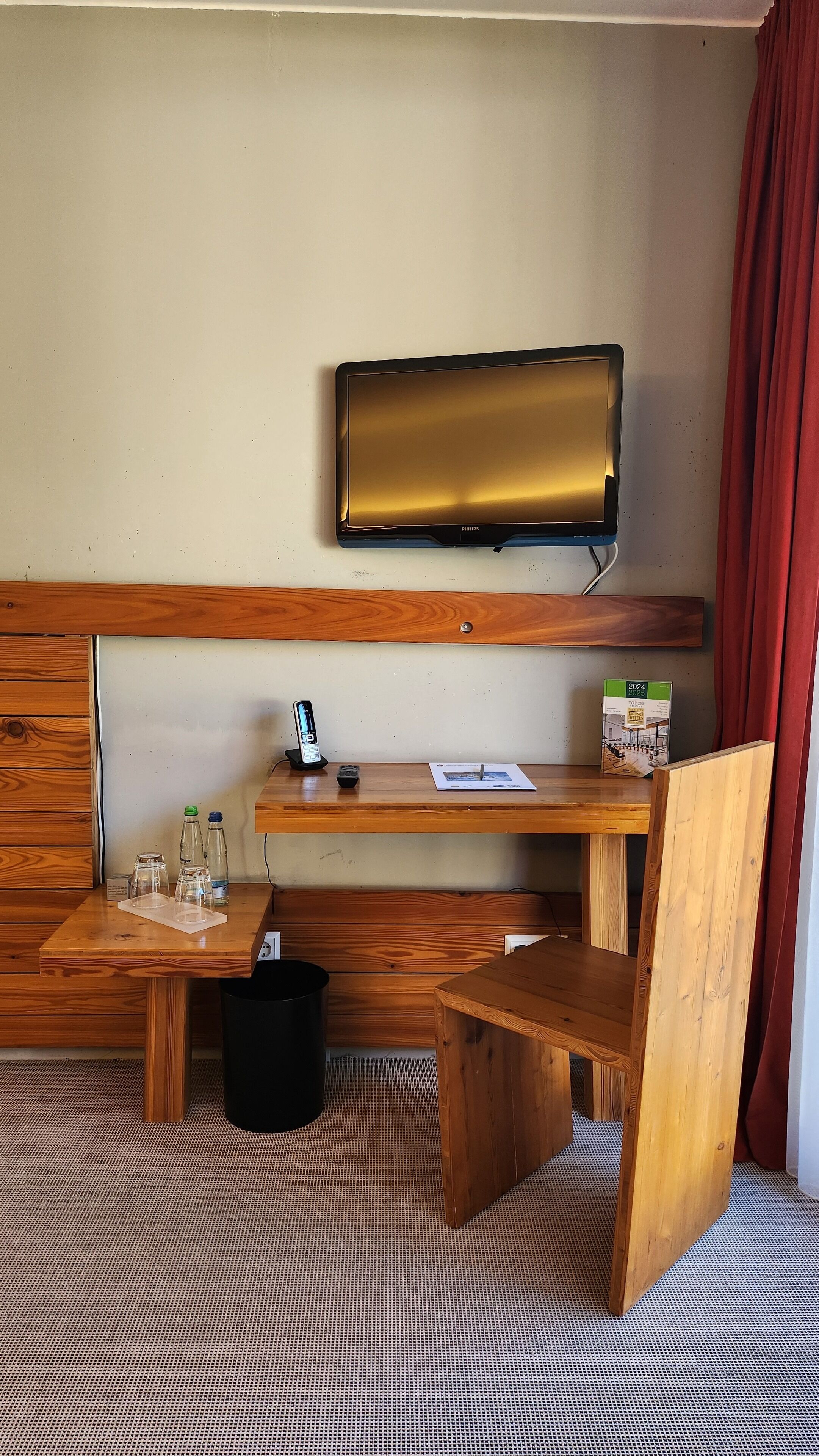 A hotel room with a wooden desk, a flat-screen TV mounted on the wall, a small trash bin, and a red curtain.