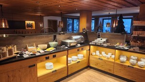 A breakfast buffet area with various food items and utensils, wooden cabinets, and a view of the outdoors through large windows. - Speidel's Brau Manufaktur (Hohenstein)