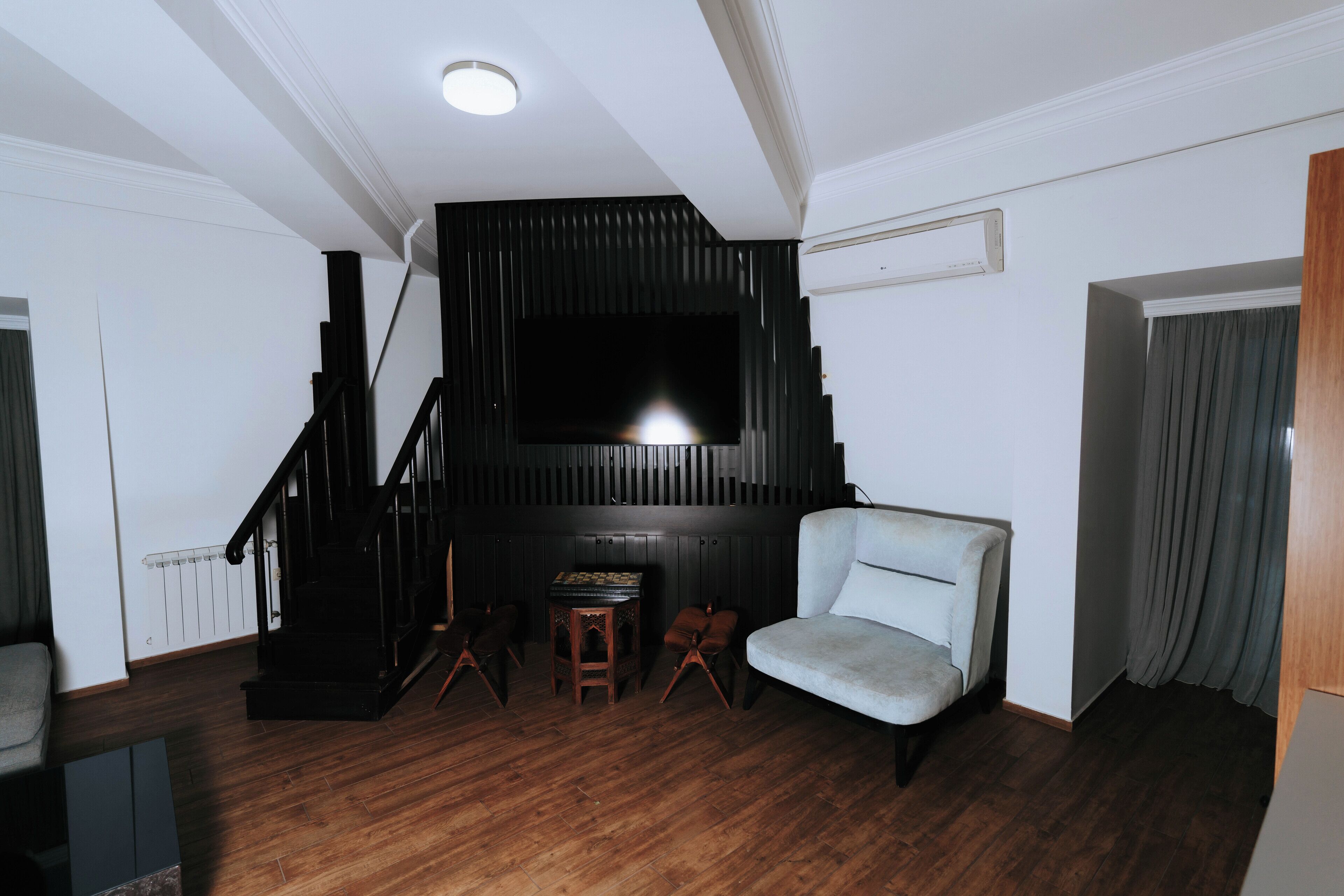 A living room with a staircase, a flat-screen TV mounted on a dark wall, a light-colored sofa, and a wooden coffee table.