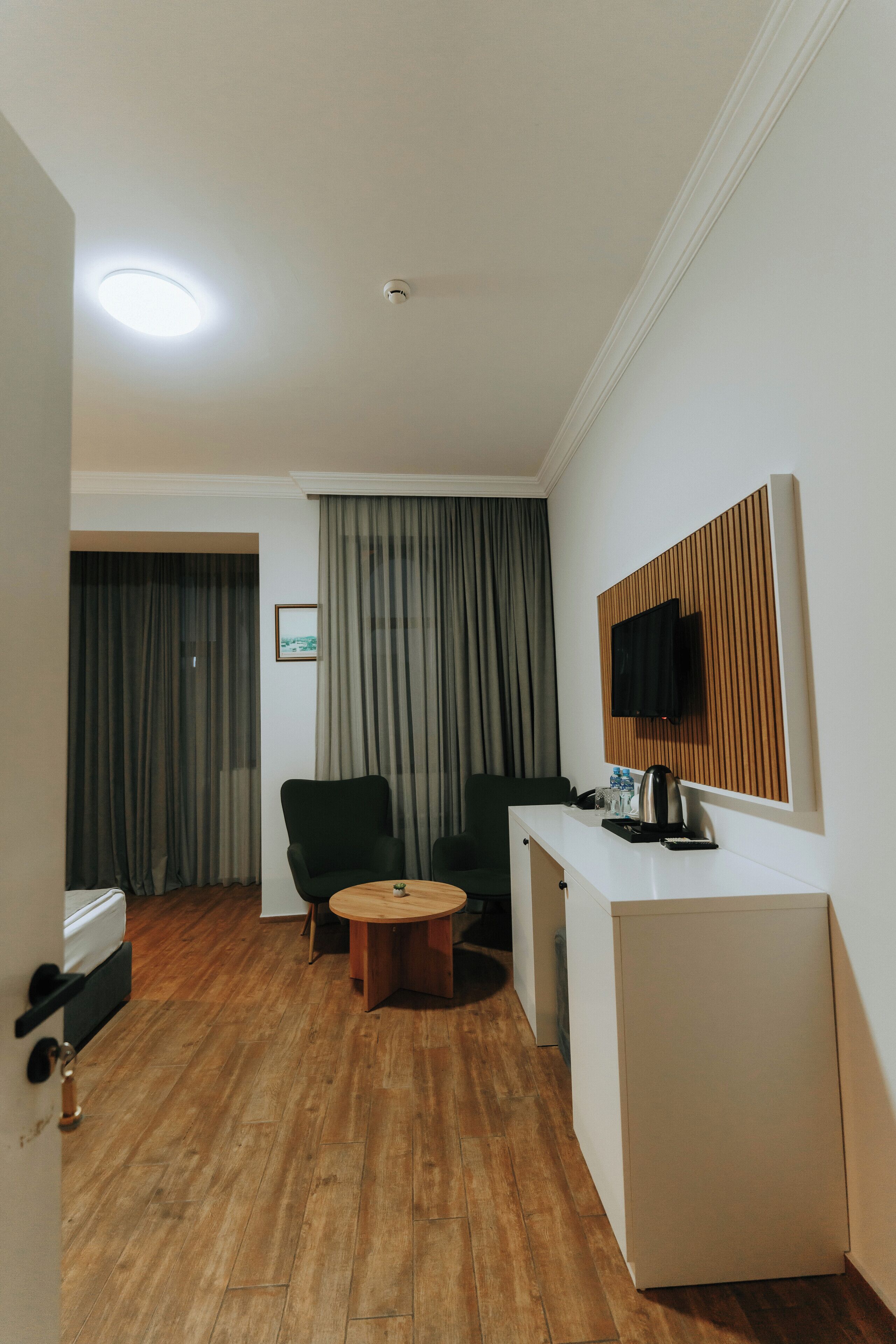 A hotel room with a wooden floor, a small round table, a green armchair, a white desk with a TV, and a window with curtains.