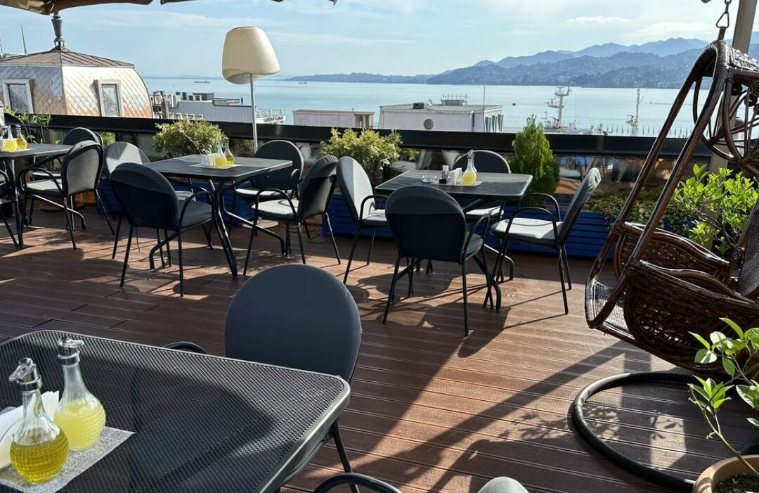 An outdoor terrace with tables and chairs, overlooking a body of water and mountains.