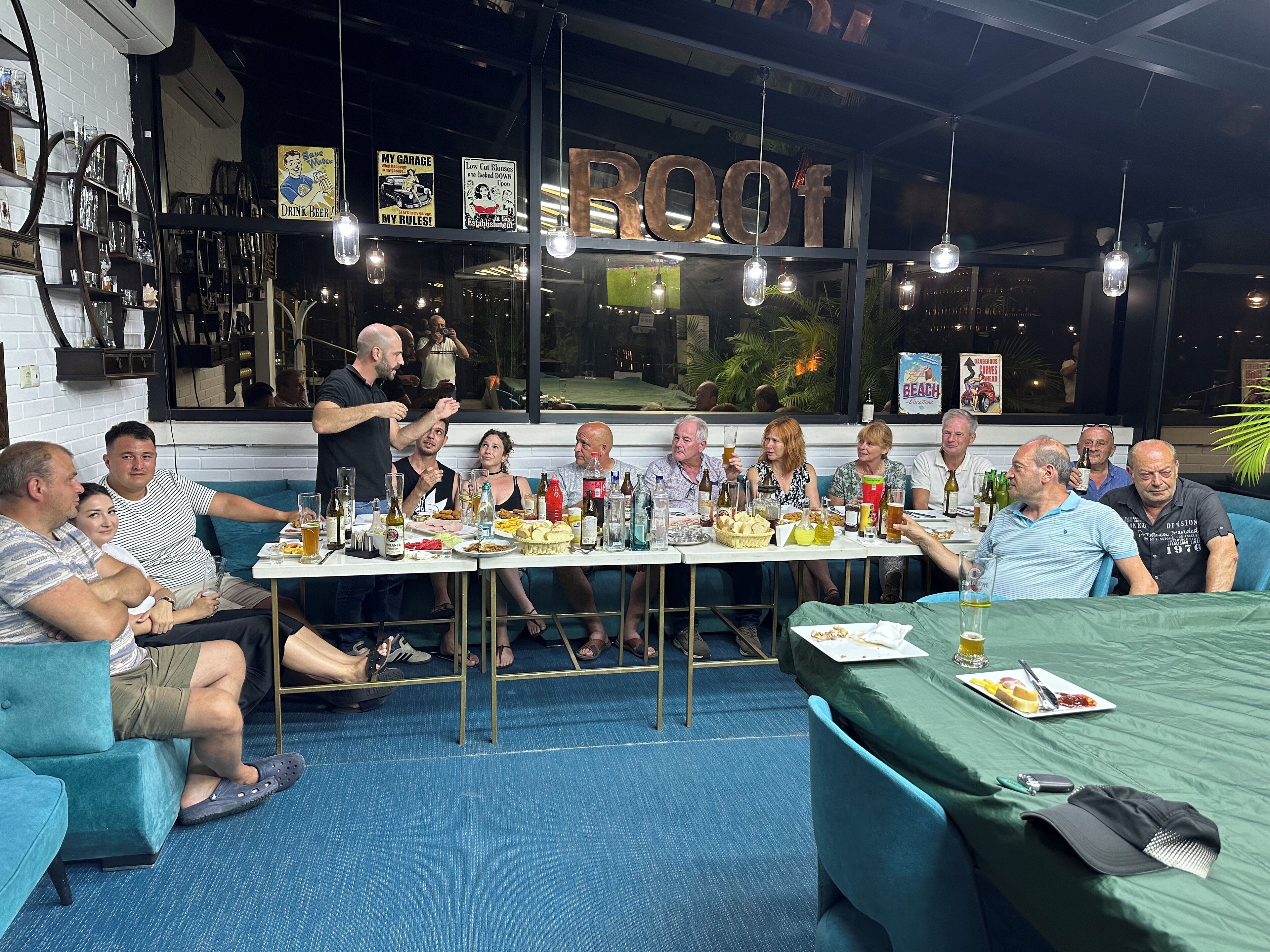 Un grupo de personas sentadas alrededor de una mesa con comida y bebidas, en una habitación con un ventanal grande y un letrero que dice 