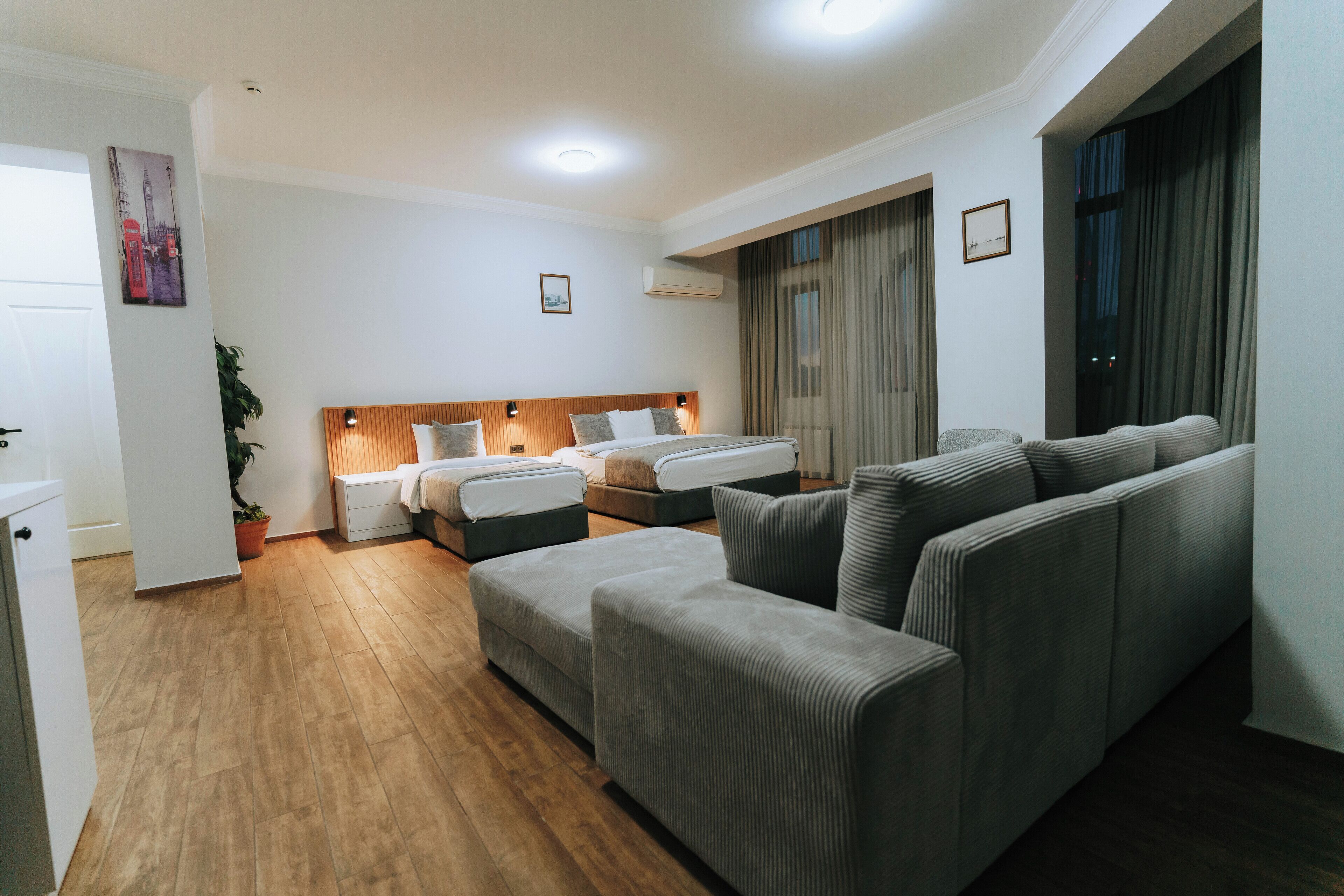 A modern hotel room with a wooden floor, a large grey sofa, a bed with a wooden headboard, and a window with curtains.