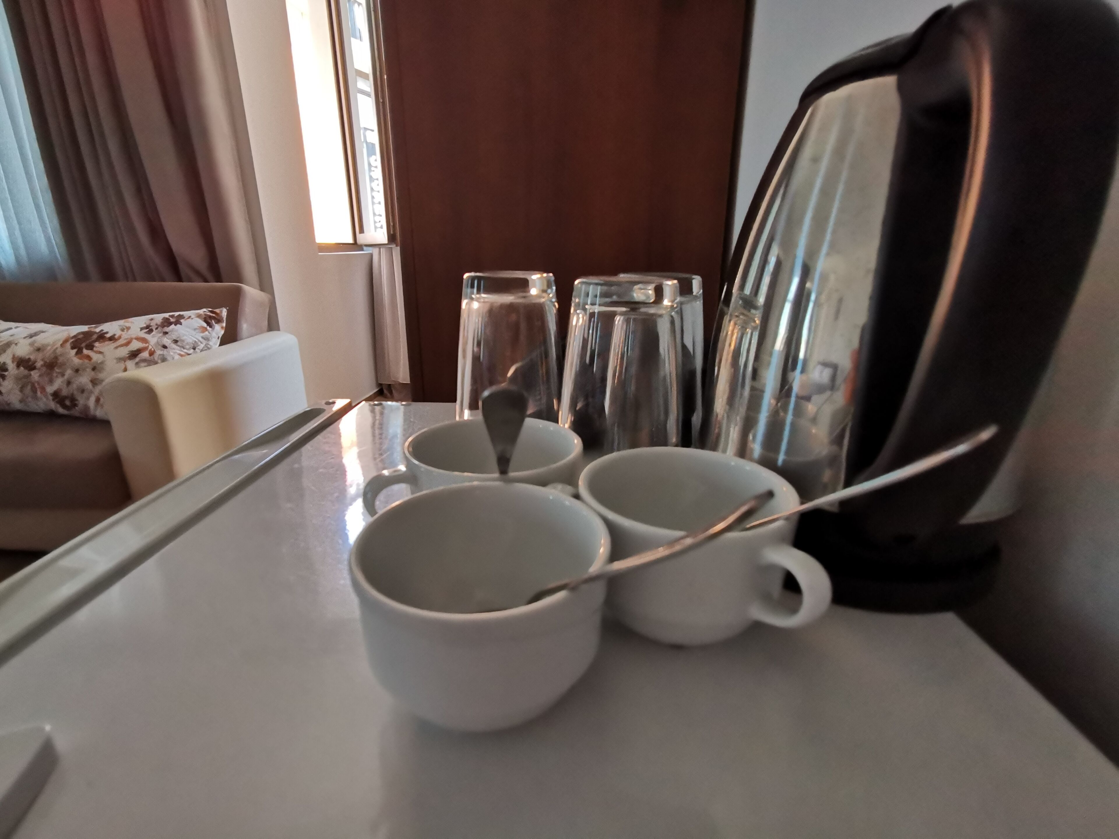 Une chambre d’hôtel avec une table à manger dressée, agrémentée de tasses blanches et d’une cuillère, un canapé avec un coussin à motifs floraux et une fenêtre avec des rideaux.