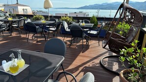 Un área de estar al aire libre con mesas y sillas, una silla colgante y vistas al mar y las montañas.