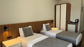 Habitación de hotel con dos camas, cabecero de madera, mesita de noche con lámpara, armario y una pequeña kitchenette.