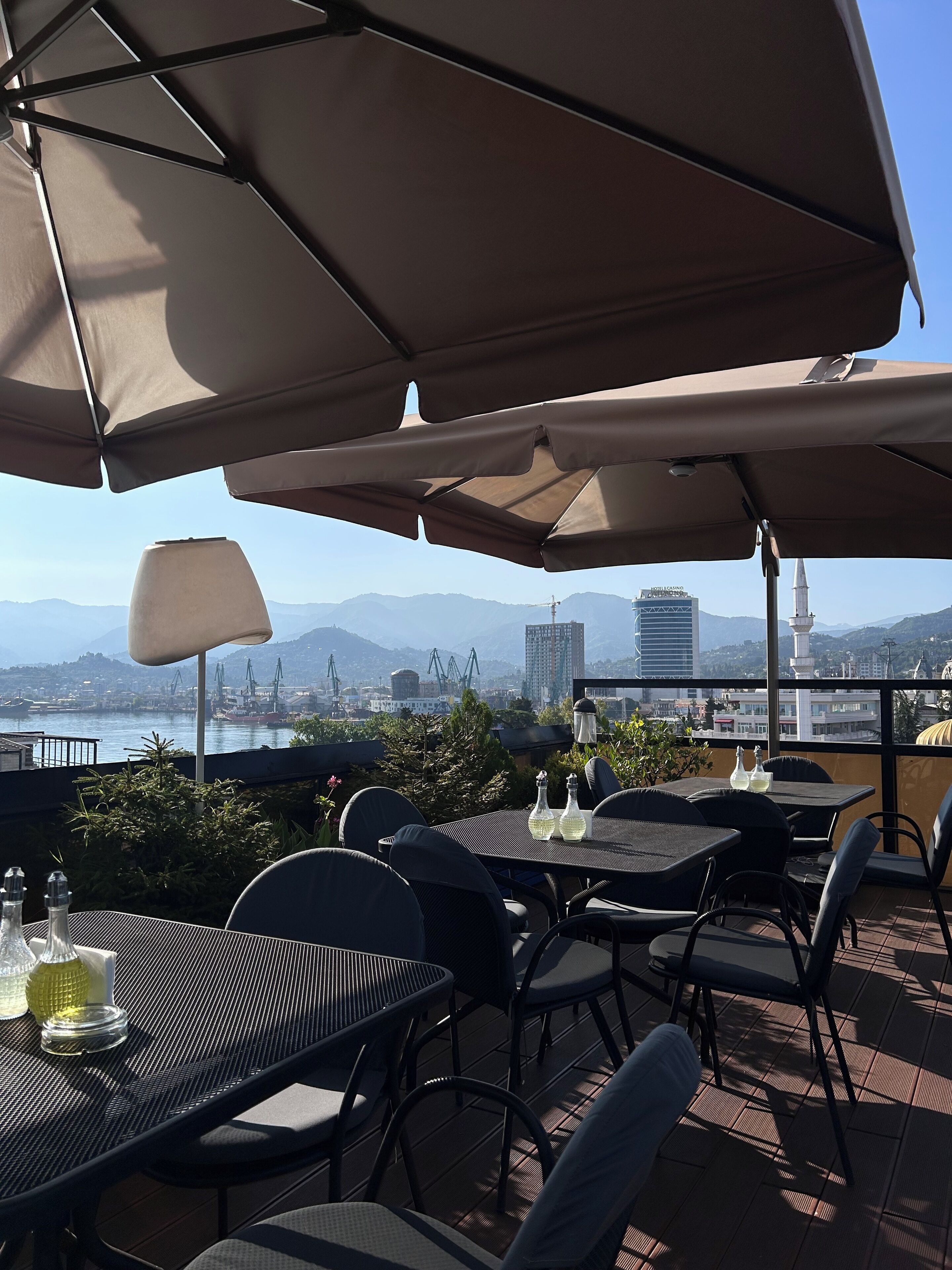 An outdoor dining area with tables and chairs, large umbrellas, and a view of a cityscape and mountains in the background.