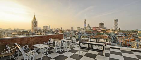 A rooftop terrace with checkered flooring, white metal furniture, and a cityscape view in the background.