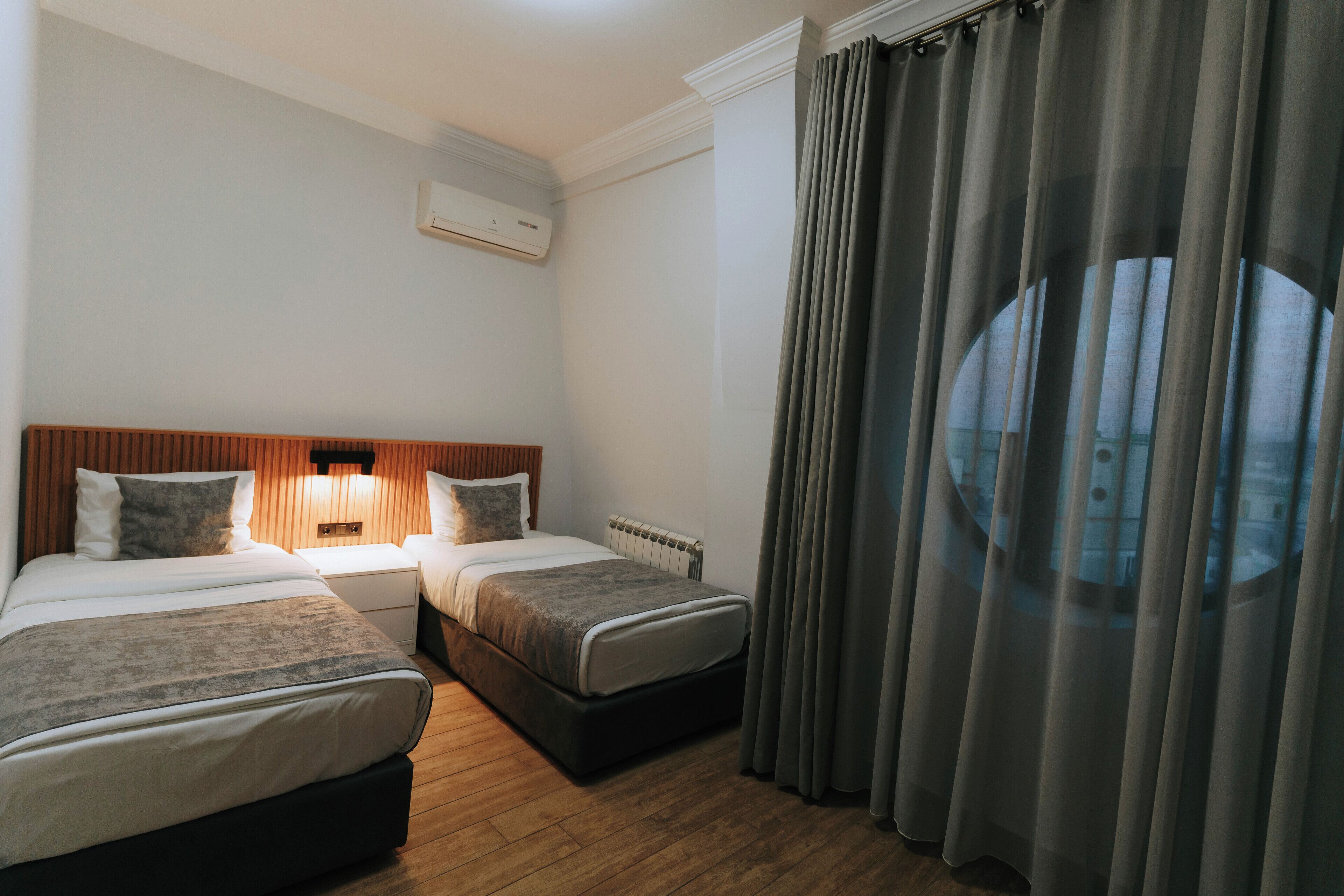 Habitación de hotel con dos camas, cabecera de madera, mesita de noche, lámpara y ventana con cortinas.