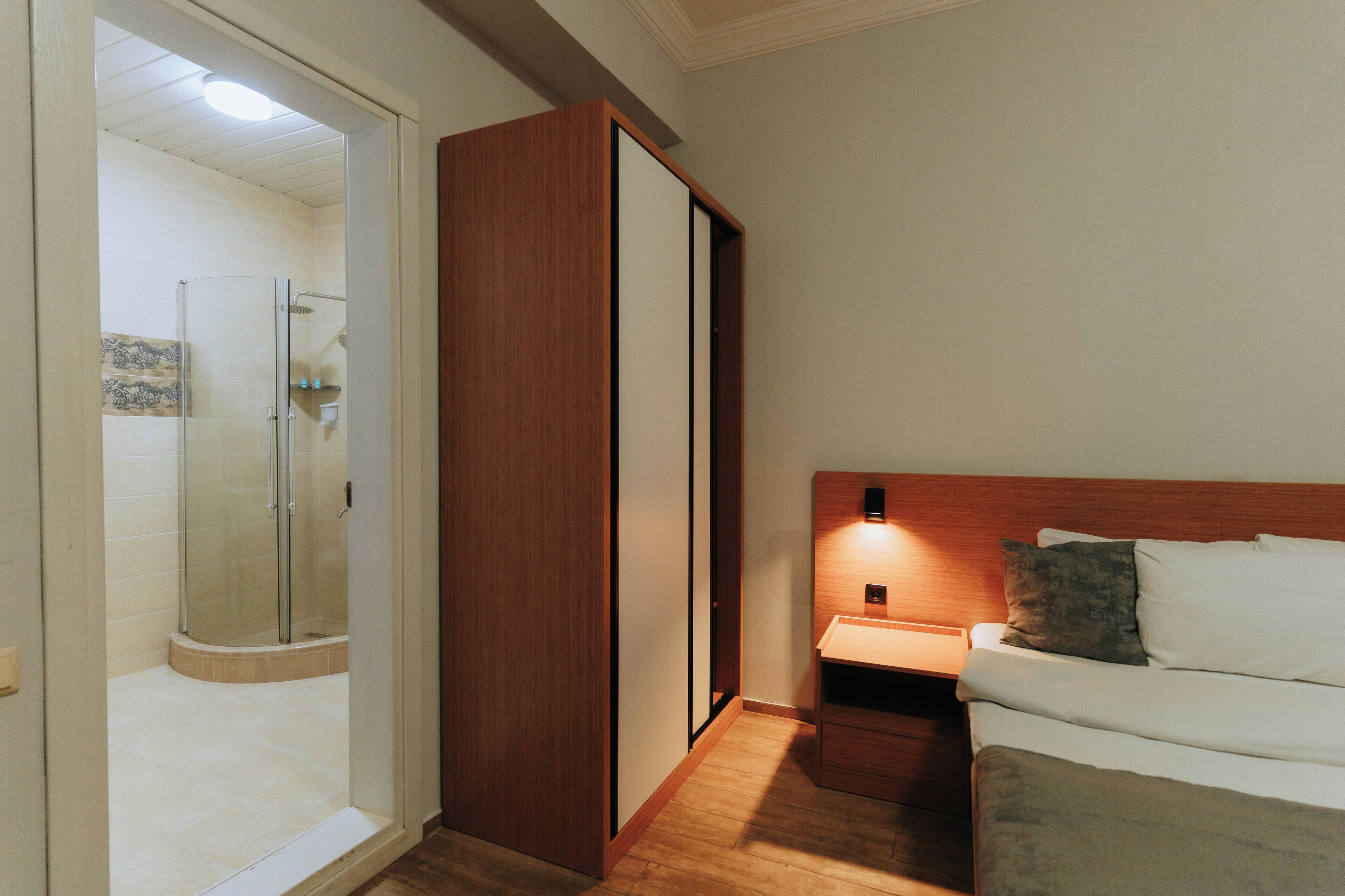 A hotel room with a wooden wardrobe, a bed with pillows, a nightstand, and a glass-enclosed shower area.