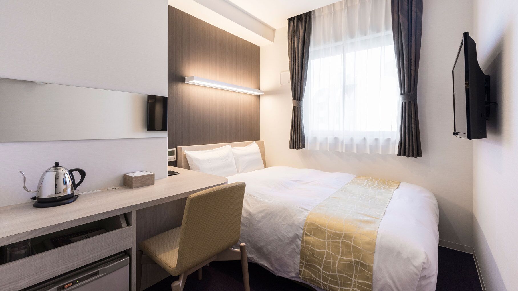 A hotel room with a bed, desk, chair, kettle, and a TV.