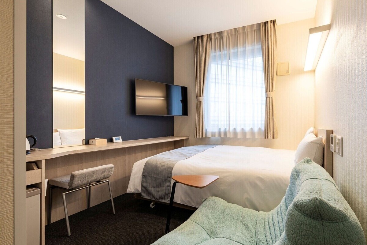 Una habitación de hotel con cama, escritorio con silla, televisor y ventana con cortinas.