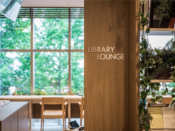 Un espace de détente dans une bibliothèque, avec un pilier en bois portant l’inscription « ESPACE DE DÉTENTE DE LA BIBLIOTHÈQUE », une fenêtre donnant sur des arbres et une étagère remplie de livres et de plantes.