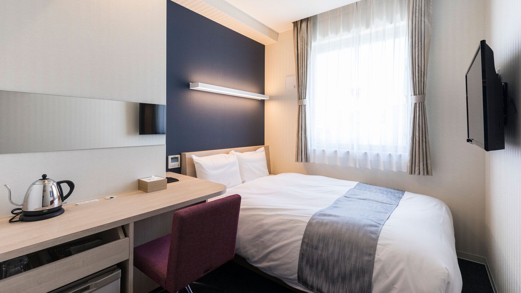 A hotel room with a bed, a desk with a kettle, a chair, a TV, and a window with curtains.