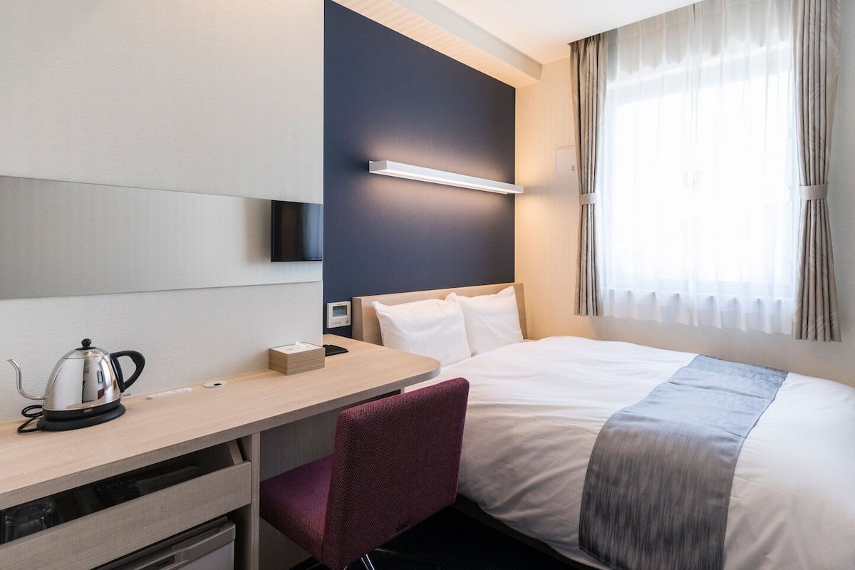 Habitación de hotel con una cama, un escritorio con hervidor, una silla, un televisor y una ventana con cortinas.