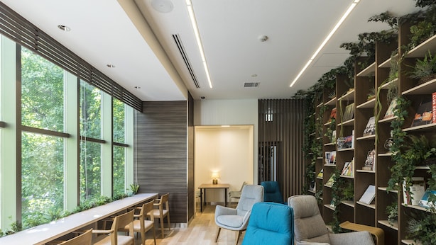 Una biblioteca moderna con mobiliario de madera, ventanas amplias y una estantería con plantas.