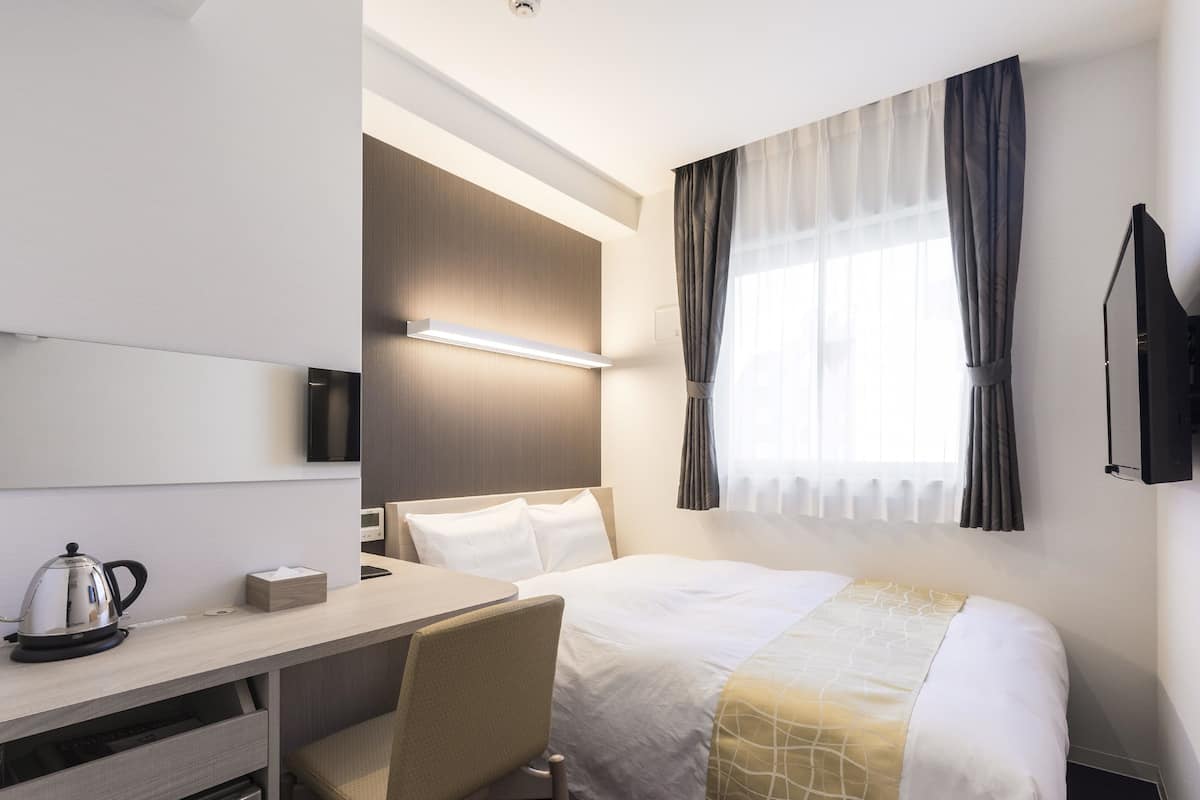Habitación de hotel con cama, escritorio, silla, televisión y ventana con cortinas.