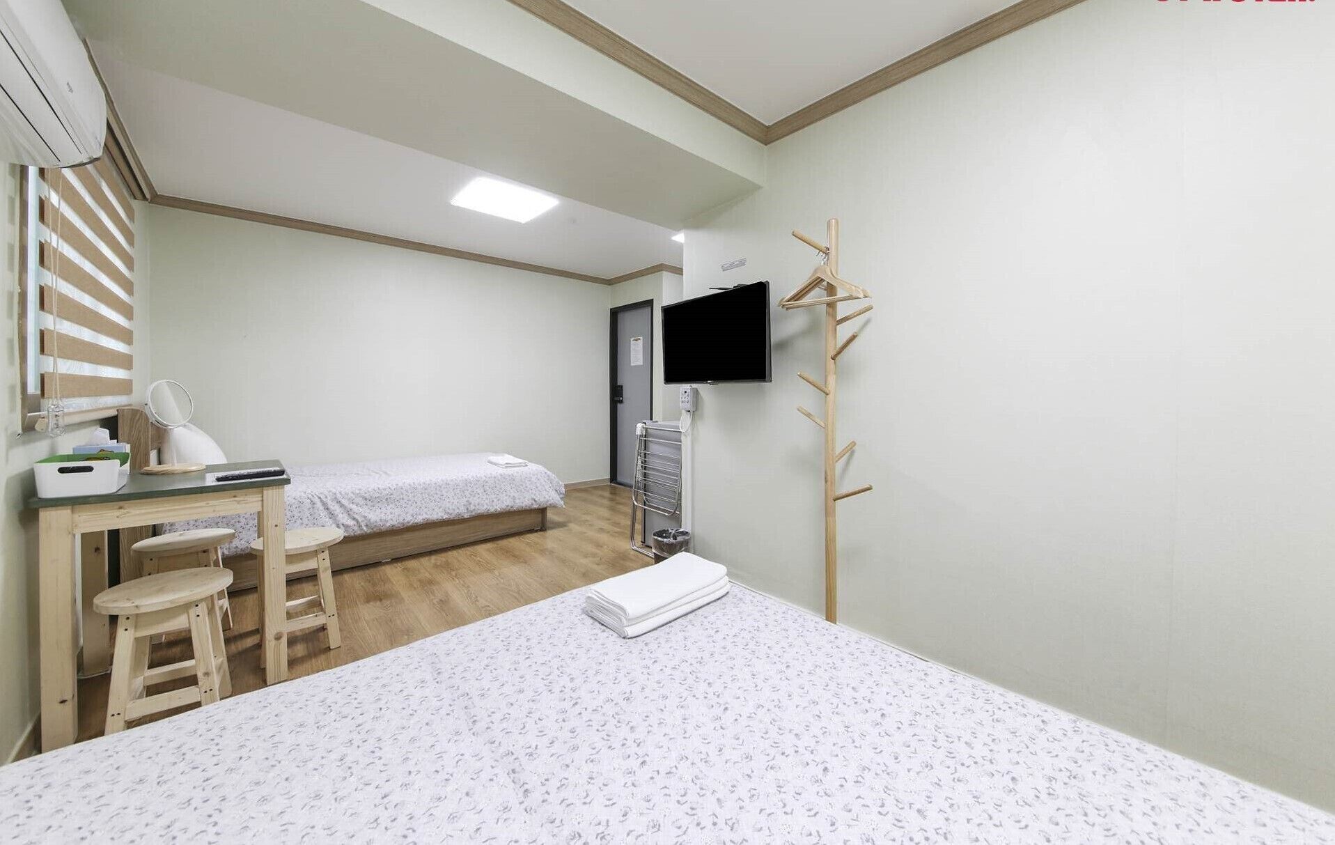 A small, clean room with a bed, a desk with a chair, a TV, and a wooden drying rack.