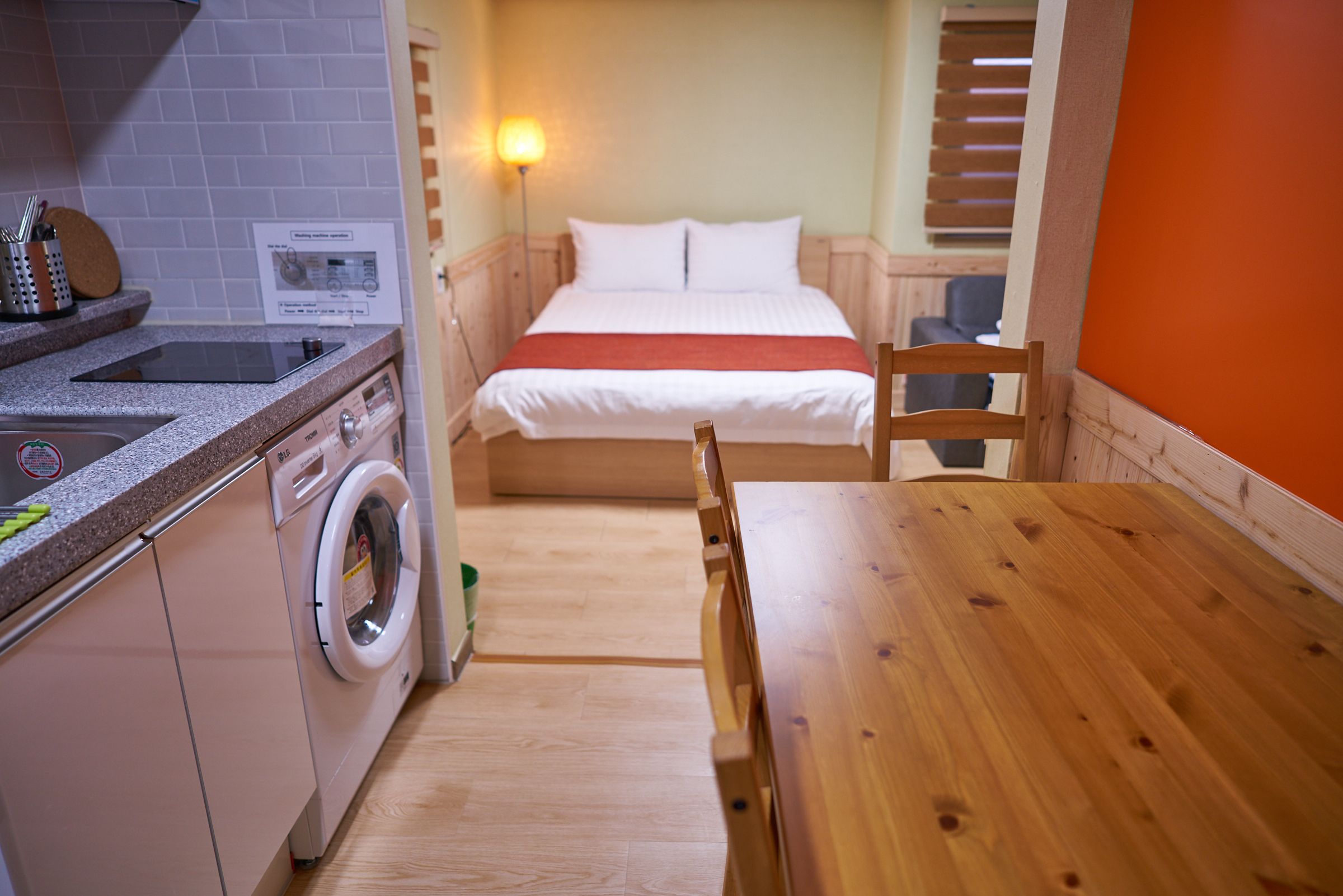 A compact hotel room with a bed, a wooden table, a washing machine, and a kitchenette.