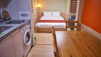 A compact hotel room with a bed, a wooden table, a washing machine, and a kitchenette.