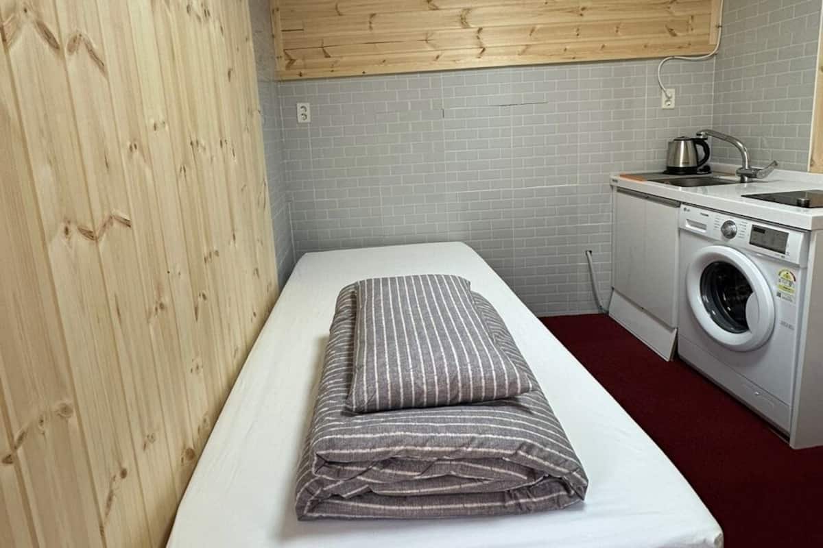 A compact room with a bed, a washing machine, and a small kitchenette.