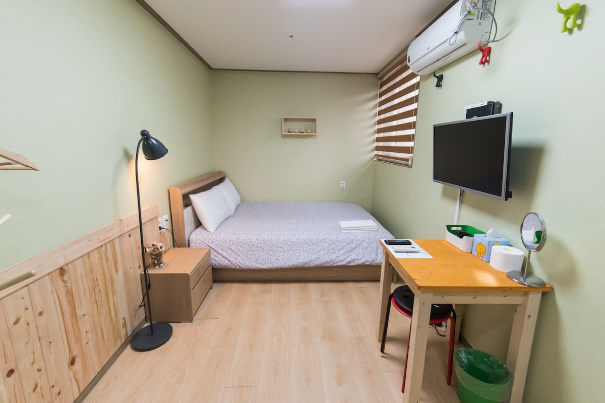 A small, well-lit room with a bed, bedside table, desk, and a TV.