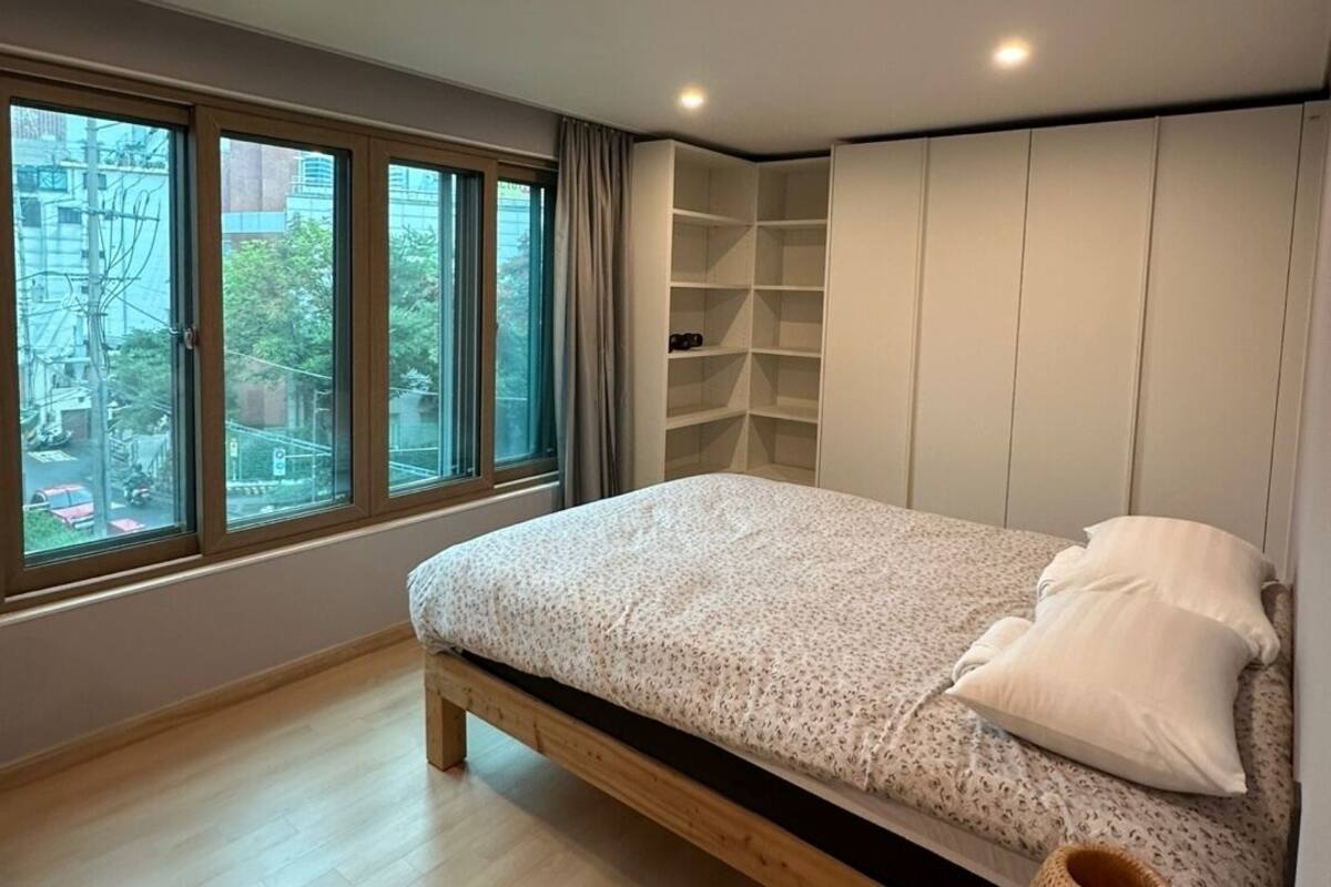 A modern bedroom with a large window, a bed, built-in shelving, and a bedside lamp.