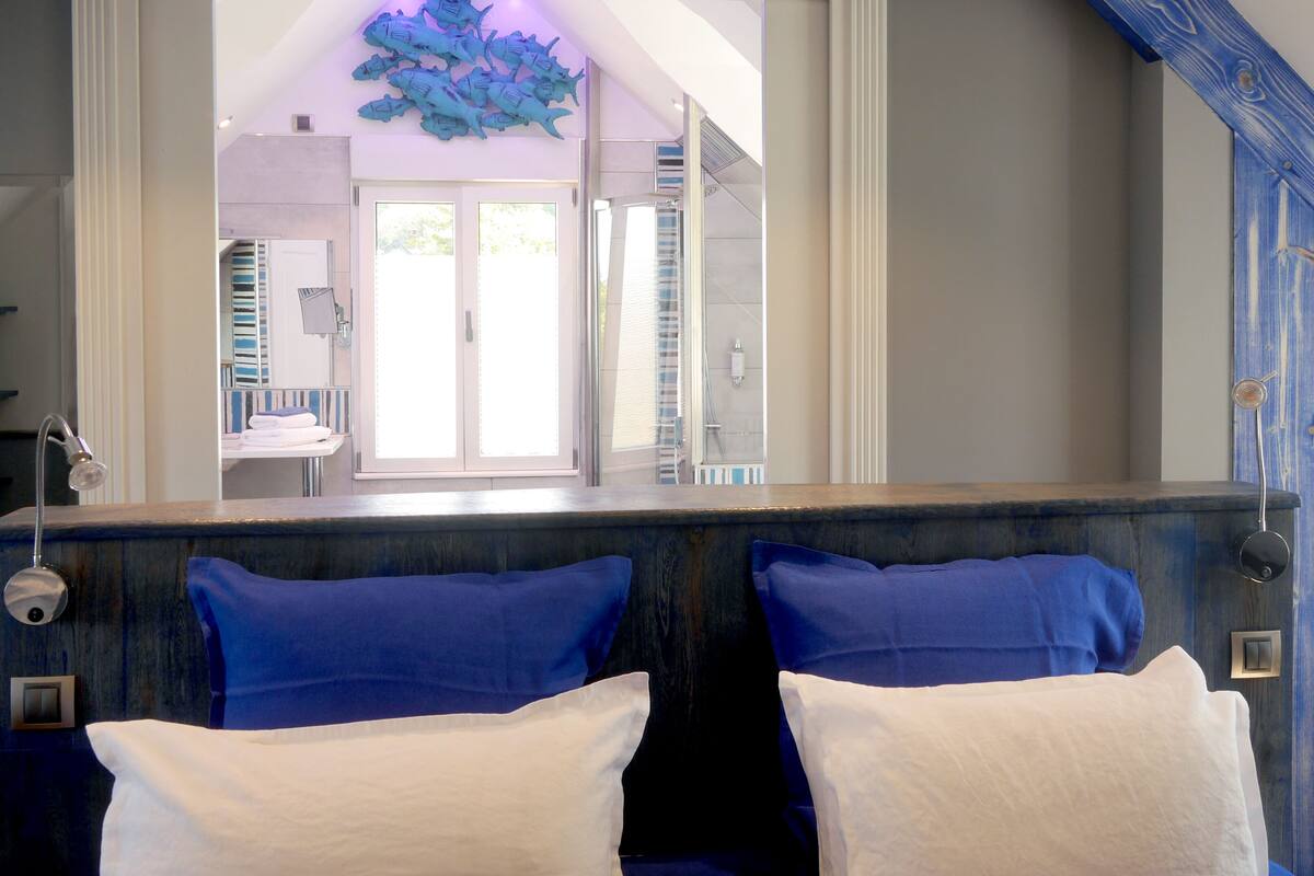 Habitación de hotel con un espejo grande, vista al baño y un cabecero de madera con almohadas azules.
