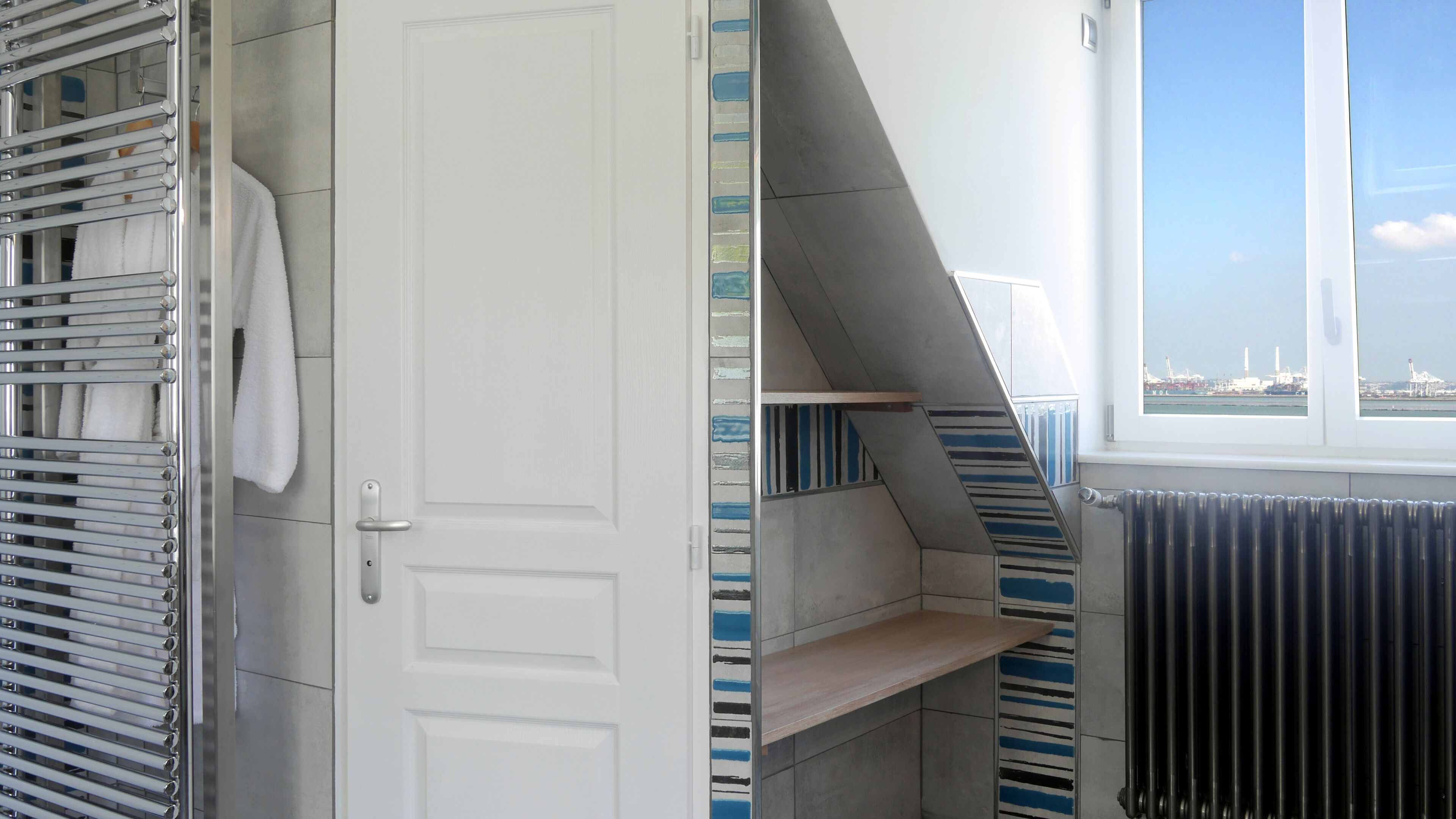 Une salle de bain moderne avec une porte blanche, un porte-serviettes, un radiateur et une vue sur un port, visible par une fenêtre.