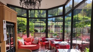 Un café soleado con mobiliario rojo, ventanas amplias y vistas a un jardín exuberante.