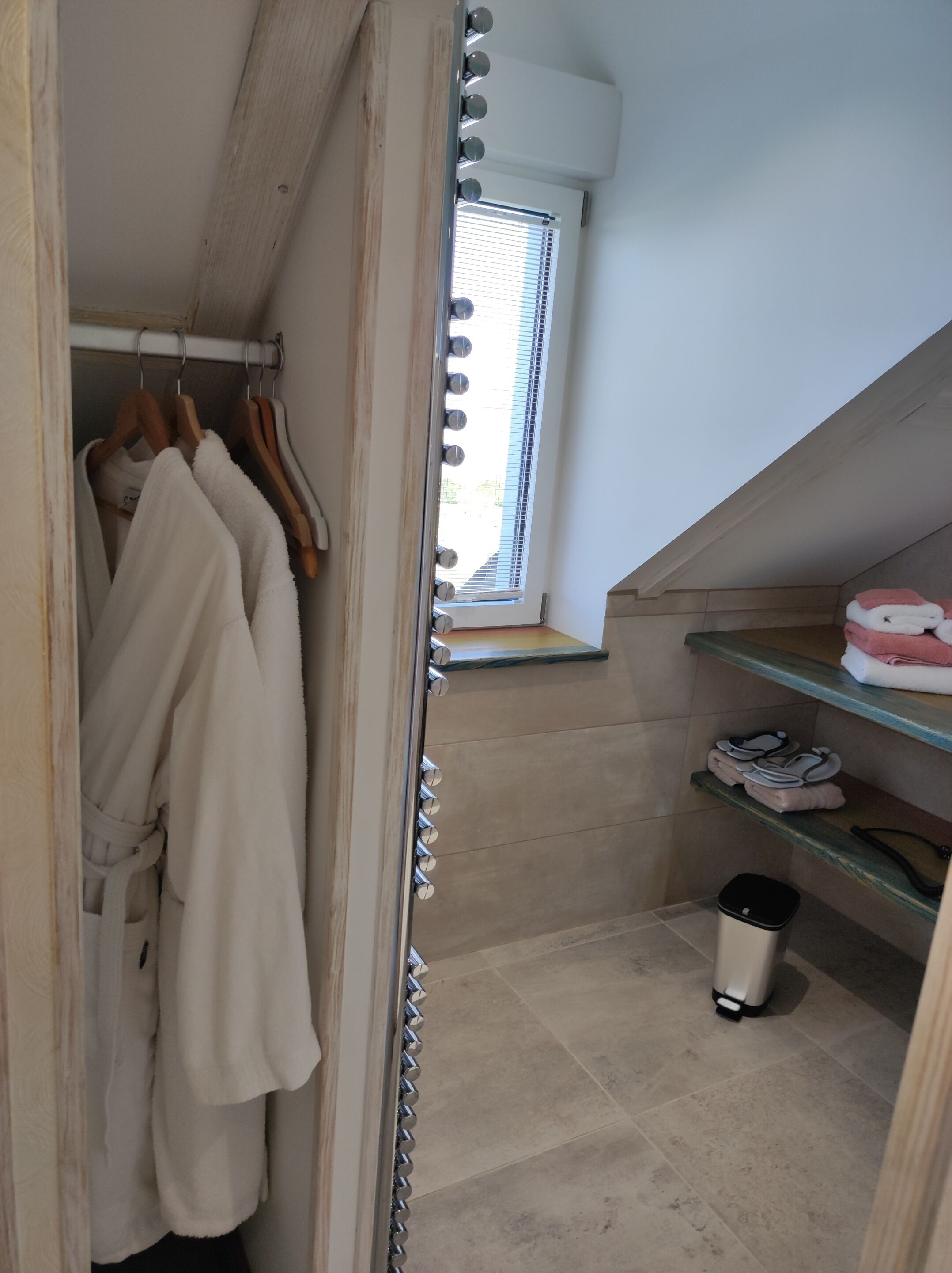 Een inloopkast met een houten deur, een raam met zonwering, en een plank met opgevouwen handdoeken en slippers.