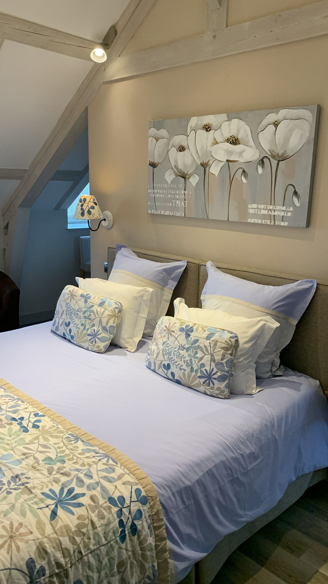 Ein Schlafzimmer mit einem großen Bett, Kissen mit Blumenmuster, einer Wandlampe und einem Blumenbild.